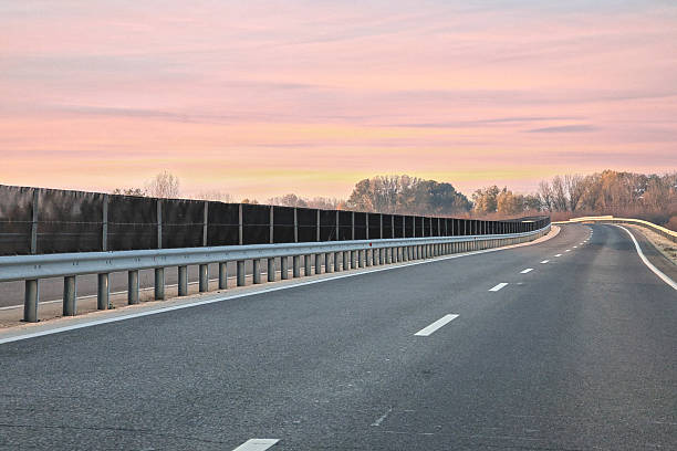 This tranquil photograph depicts an empty asphalt road curving gently to the right, marked by crisp white lane dividers and a solid white shoulder line. The road is bordered by a metallic silver guardrail with a line of autumnal trees visible behind the barrier, all set beneath a pale, soft pastel pink and orange sky. This image is excellent for symbolizing travel, quiet journeys, road infrastructure, construction, solitude, planning, or the smooth transition of a new day or ending day.