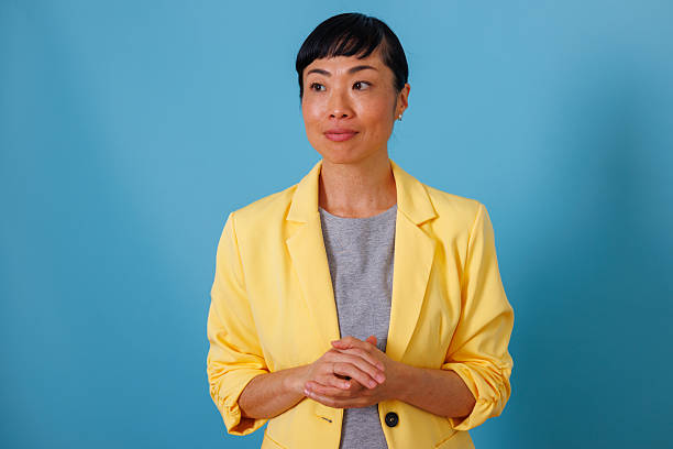 istock Professional asian woman in yellow blazer posing with hands clasped, thinking on blue background 2246495116