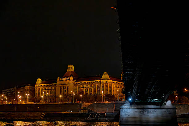 Budapest, Hungary - May, 2025Hungarian Building stands prominently along the Danube River at night, its ornate facade illuminated by warm golden lights that reflect on the dark water surface. The neo-Gothic structure features multiple towers, spires, and arched windows, with the central dome visible. Streetlights and bridge structures line the riverbank, casting additional glows.