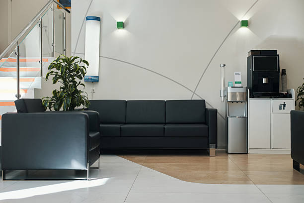 Empty modern waiting area showing black leather sofas, coffee machine, water dispenser and potted plant, clean tiled floor and staircase with glass railing in background