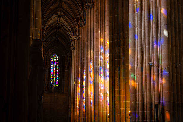 Inside the quiet sanctuary of Batalha Monastery, vivid reflections from intricate stained glass windows cast a kaleidoscope of colors across the ribbed stone columns and walls. The warm, ambient lighting contrasts with the dark Gothic recesses of the architecture, creating a mesmerizing atmosphere of stillness and spiritual reverence. A shadowed statue watches over the sacred space, enhancing the mood of timeless devotion and artistic craftsmanship within this UNESCO-listed Gothic masterpiece.