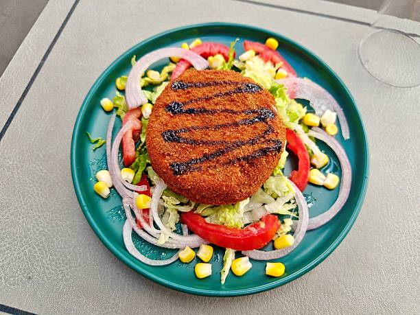 A golden-brown, breaded and grilled Camembert cheese patty rests on a vibrant bed of fresh lettuce, red onion rings, sweet corn kernels, and juicy tomato slices. A delightful meal served in Annaba, Algeria.