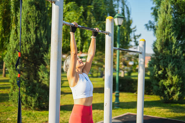 Active mature woman working out outdoors, performing a pull-up exercise on a metal bar in a sunny park. She is wearing coral leggings, a white sleeveless crop top, and fitness gloves. The image represents strength, health, motivation, and active lifestyle through outdoor bodyweight training.