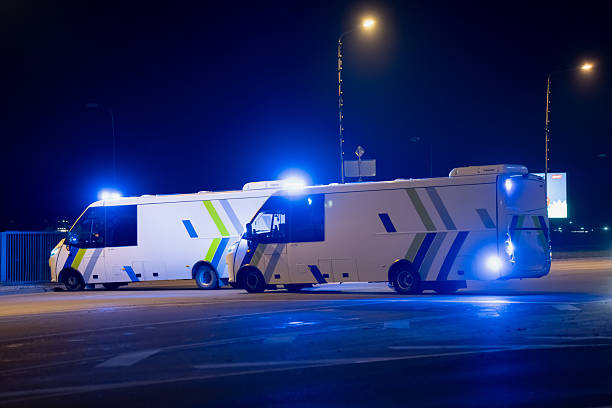 Two police response vehicles with active blue lights positioned on an empty roadway at night, showing reflective markings, operational stance and the distinct silhouette of mobile command units in a controlled area