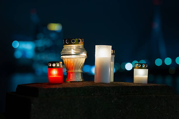 Collection of lit lantern candles on a Latvian waterfront structure, framed by distant architectural lights and soft bokeh from a nearby bridge, forming a peaceful night scene filled with warm reflective illumination