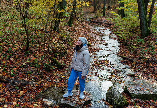 A lone woman stands on a stone in a shallow autumn stream, surrounded by fallen leaves and warm forest colors. The moment captures peaceful outdoor exploration, seasonal mood, and connection with nature during a calm fall walk.