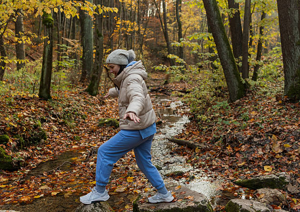 A woman carefully steps across stones as she crosses a small forest stream covered with golden leaves. The scene reflects autumn exploration, balance, and the beauty of seasonal outdoor activity.