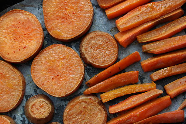 Baked sweet potato rounds and carrot sticks seasoned with herbs and spices on dark baking tray. Roasted vegetables showing caramelized edges and golden color after oven cooking with visible seasoning