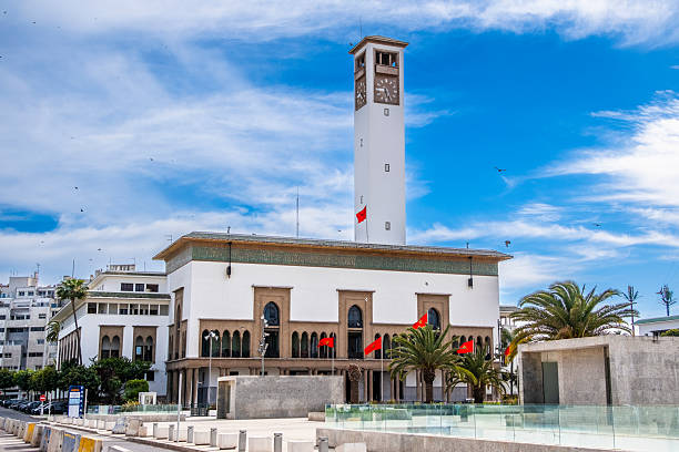 Casablanca, Morocco â View of the iconic Casablanca City Hall (HÃ´tel de Ville) with its prominent clock tower, Moroccan flags, and palm trees under a vibrant blue sky. This historic building, located at Mohammed V Square, showcases a blend of traditional Moroccan and Art Deco architectural styles. A symbol of civic pride and colonial-era heritage, it represents the cultural and administrative heart of the city.