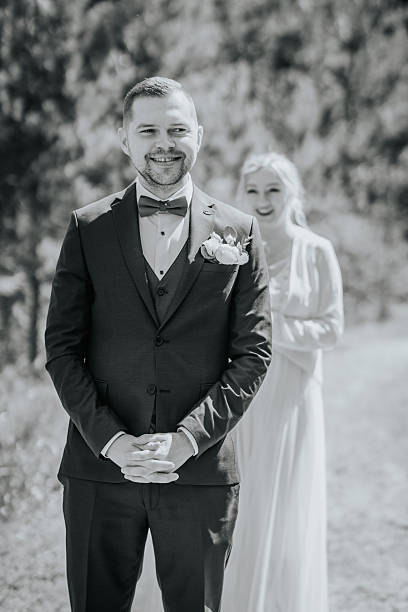 istock Groom in formal black suit with bow tie stands outdoors, smiling joyfully as bride approaches in elegant white dress, capturing a romantic wedding moment in nature 2246540235