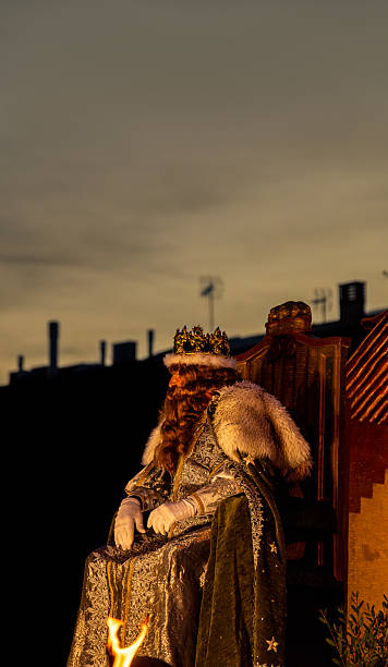 King Melchior, one of the Three Wise Men, sits on a throne atop a float during the Three Kings Parade on a street at dusk.