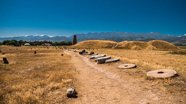 The Burana Tower is an ancient brick minaret standing alone on a wide plain, surrounded by historic ruins and mountain views.