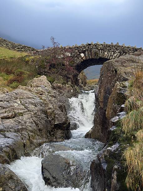 An arched stone footbridge crosses over the River Derwent near to Saithwaite on a hiking trail in the Lake District on a moody day