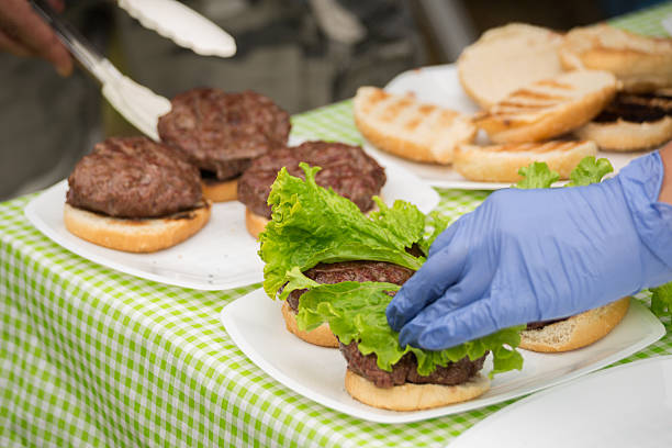 Close-up of a gloved hand placing fresh green leaf lettuce onto beef patties on toasted burger buns. Several open-faced burgers sit on white plates over a green gingham tablecloth, with grilled bun tops and tongs visible in the background, suggesting a picnic, BBQ, or catered event.