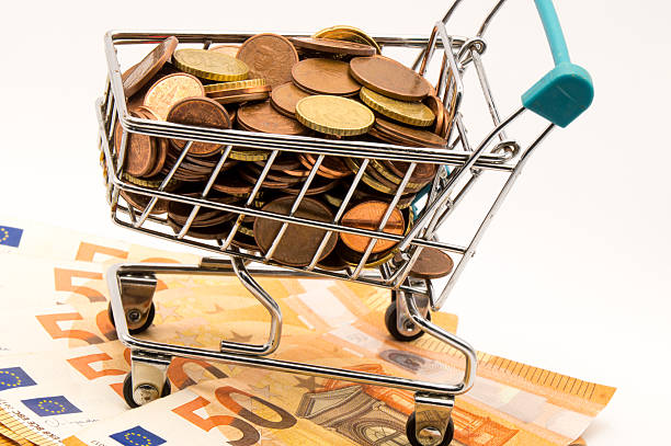 Descripción: A dynamic, diagonal close-up of a mini shopping cart loaded with Euro coins and resting on a foundation of €50 banknotes. This creates visual interest for topics like market risk, financial leverage, and money flow.