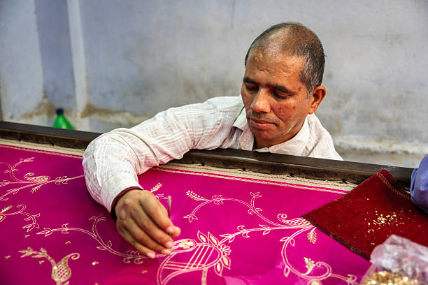 An artisan leans over a workbench, carefully applying gold embellishments and embroidered details to a vivid magenta textile. The close-up shows delicate floral and vine motifs being worked with a small tool; a red cloth holding sequins and a small bag of metallic pieces sit nearby. The setting appears to be a modest workshop where precise handcrafting is taking place.