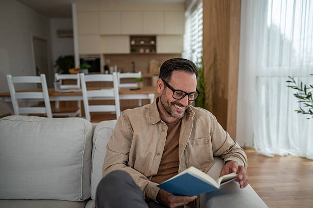 istock Smiling man relaxing on couch reading book at home 2246570385