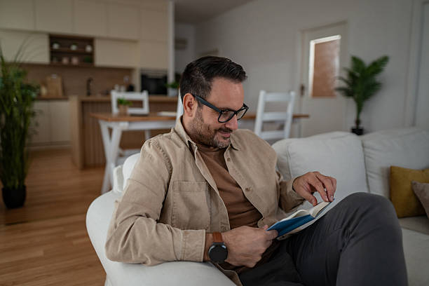 istock Man relaxing on couch reading book at home 2246570389