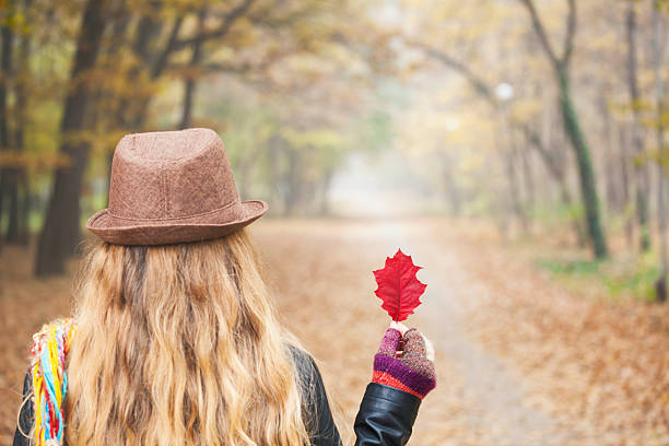 Rear view of a woman standing on a leaf-covered path in a misty forest, holding a red leaf. Concept of personal choice, individuality, originality.