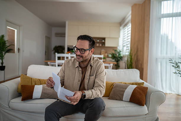 istock Smiling man opening letter on living room sofa 2246571950