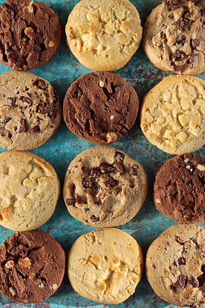 istock Full frame image of rounds of chocolate chip shortbread biscuits flat lay, rows of alternating triple chocolate chip, milk chocolate chip and white chocolate chip cookies, mottled blue-green background, elevated view 2246585897