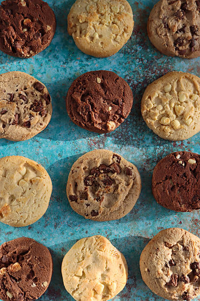 istock Full frame image of rounds of chocolate chip shortbread biscuits flat lay, rows of alternating triple chocolate chip, milk chocolate chip and white chocolate chip cookies, mottled blue-green background, elevated view 2246585935