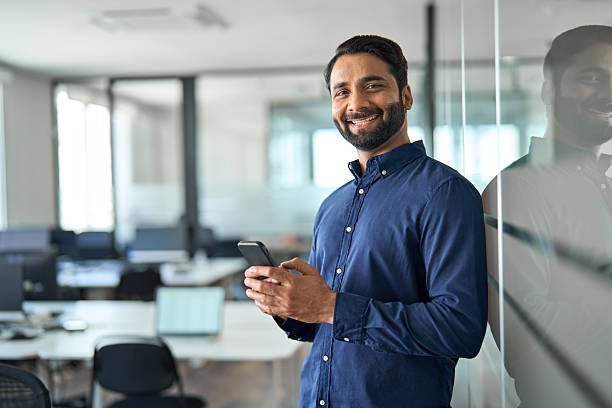 istock Smiling Indian businessman using mobile cell phone working standing in office. 2246601238