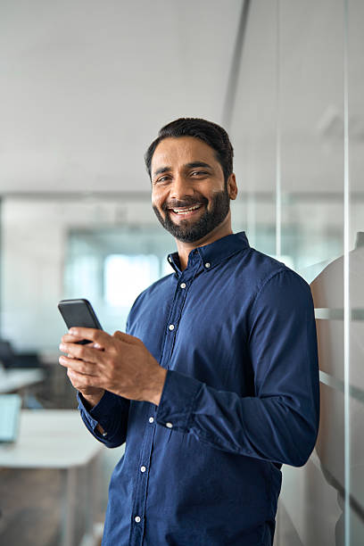 istock Smiling Indian businessman using mobile cell phone standing in office. Vertical 2246601243