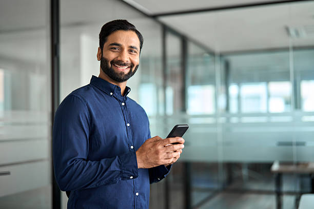 Happy Indian business man using cell phone looking at camera standing in office.