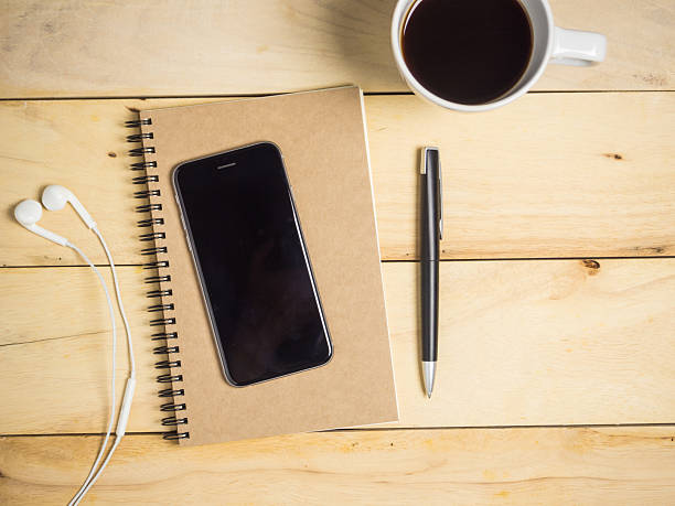Blank brown notebook with smartphone, earphone, pen & coffee on wooden table.
