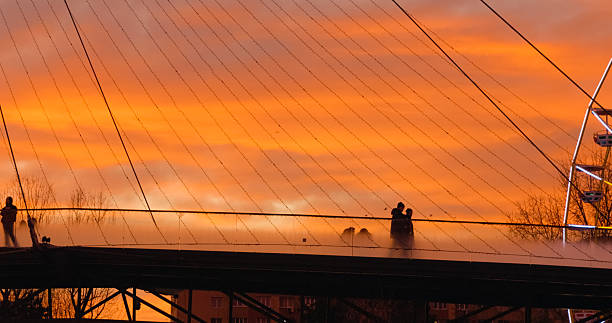 Silhouetted individuals traversing transparent pedestrian bridge during golden hour sunset, capturing urban architectural elegance against dramatic orange sky