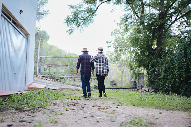 Mature couple walking hand in hand along a rural dirt path by a gate, enjoying a peaceful countryside stroll amid green trees and warm, relaxed togetherness