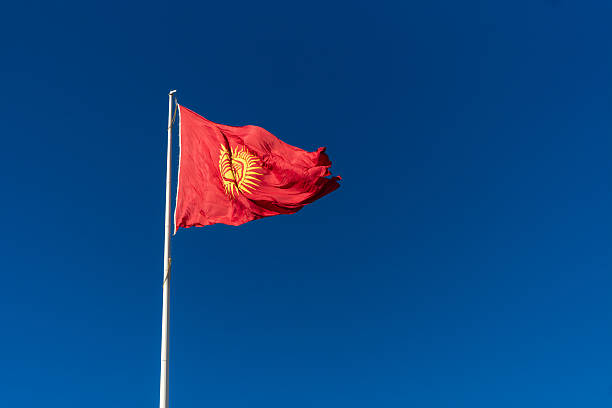 istock Red banner featuring a central golden sun with forty rays and a tunduk pattern, representing unity, heritage, and the nomadic culture of Kyrgyzstan, fluttering prominently in open daylight. 2246646456