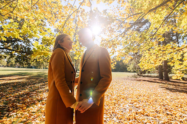 A mature couple enjoys a peaceful moment together in a park during autumn, surrounded by warm fall colors. They stand closely to each other, smiling and talking, with golden leaves scattered on the ground and trees glowing in seasonal hues