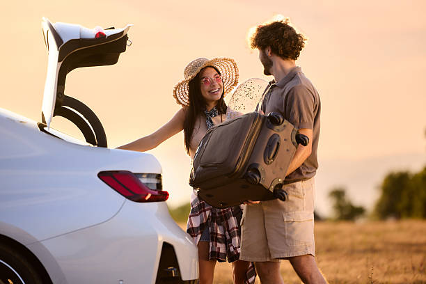 Happy couple communicating while loading their suitcase into a car trunk before a road trip at sunset. Copy space.