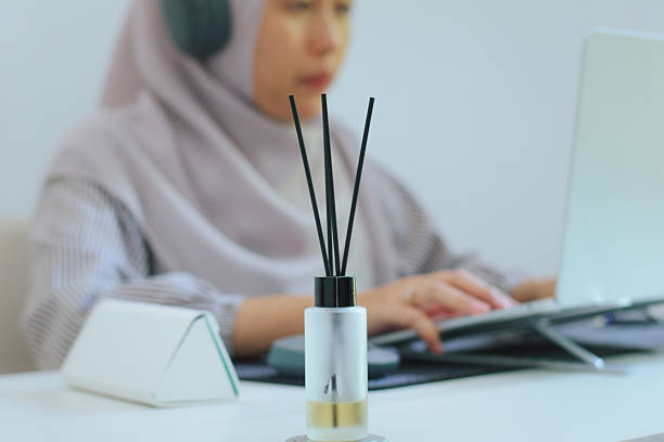 Close-up detail shot of a reed diffuser on a clean white home office desk with  woman is working on her laptop on the background. Concepts of comfort, wellness, ambiance, and self-care within the context of remote work, WFH, or modern living spaces