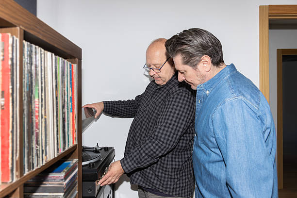 Two mature men looking at a turntable as one of them plays a vinyl record. Realistic and warm home scene, horizontal shot with copy space. Concept of friendship and music lifestyle