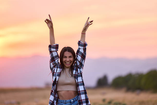 Portrait of young cheerful woman having fun with her arms raised during springtime at sunset and looking at camera. Copy space.