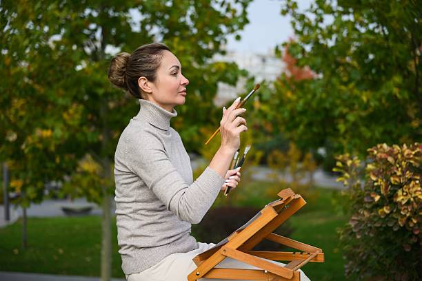 Woman painting outdoors in a park, holding a brush in front of her.