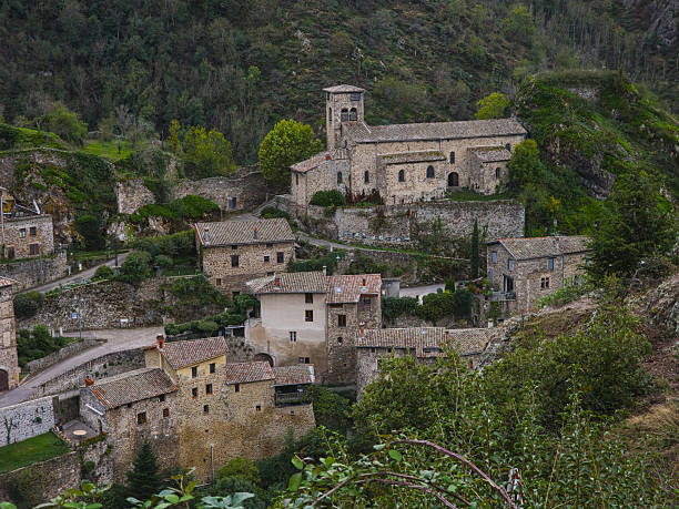 Editorial image of the historic Saint-Jean-Baptiste church and surrounding village buildings in Malleval, France, taken on October 4, 2025. The photograph captures a scenic view of the medieval architecture built into the hillside, with stone walls, tiled roofs, and lush vegetation.
