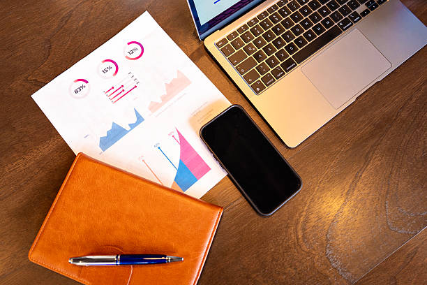 Close-up of a planner, smartphone, and business reports on a wooden desk next to an open laptop. Ideal for topics related to productivity, hybrid work, organization, digital workflow, financial analysis, and modern office routines. Keywords: office, planner, notebook, smartphone, business reports, analysis, productivity, hybrid work, desk, laptop.