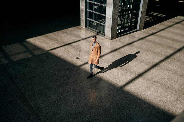 A mature man in stylish business attire walks into a subway station, photographed from a distance. His long shadow stretches behind him, capturing a dramatic urban atmosphere and a sense of movement in the city