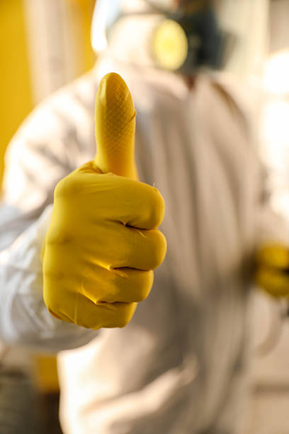 Worker In A Protective Coverall, Face Shield And Respirator Uses A Backpack Sprayer To Apply Disinfectant Along Windows And Radiator In A Sunlit Corridor.