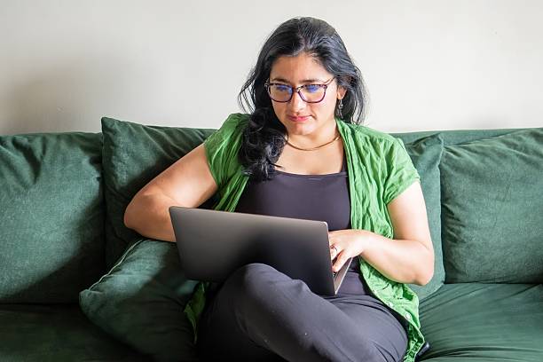 A woman is sitting on a couch and using a laptop. She is wearing glasses and a green jacket