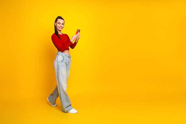 istock Young woman in red top and jeans smiles while using a smartphone against a bright yellow background for lifestyle fashion advertising and shopping promotion 2246683406