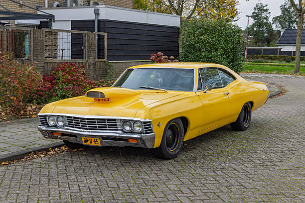 A vibrant yellow classic American muscle car, a 1967 Chevrolet Impala, parked on a paved residential street. Retro automotive. Utrecht Netherlands, 29 October 2025.