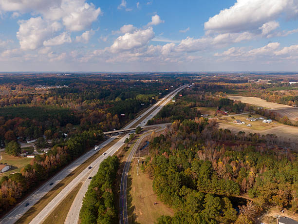 A comprehensive collection of drone aerial photographs capturing every aspect of the critical junction where Interstate 40 (I-40) meets Interstate 95 (I-95) near Benson, North Carolina. Also featured are high-altitude and top-down (nadir) views that showcase the complex geometric patterns of the multi-level overpasses, ramps, asphalt lanes, and traffic flow, emphasizing civil engineering, infrastructure, and the major transportation arteries of the US East Coast. The surrounding landscape features a mix of rural and suburban autumnal terrain, including a small pond adjacent to the looping ramps.
