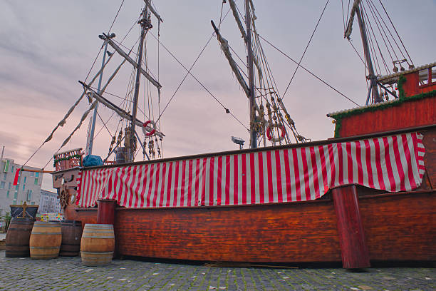 A historic ship is moored at the waterfront, showcasing a striped sail and wooden barrels during sunset.