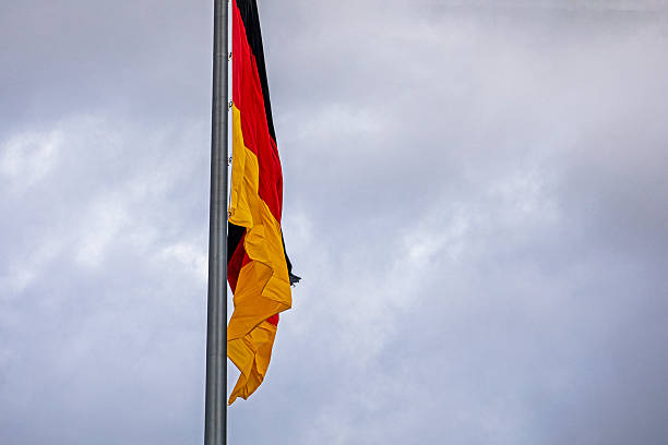 The German national flag, featuring black, red, and gold horizontal stripes, waves majestically against a clear blue sky with scattered clouds, symbolizing unity, sovereignty, and national pride.