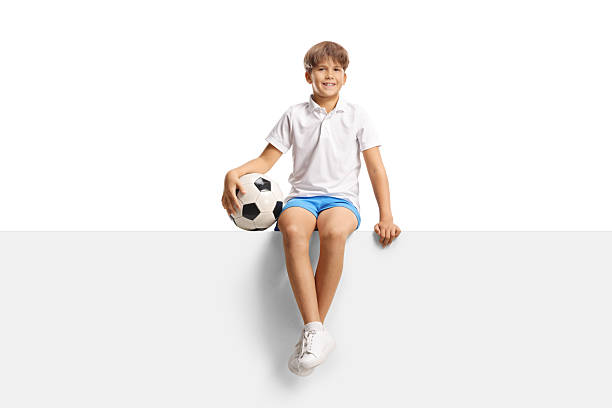 istock Happy kid holding a football and sitting on a blank panel 2246719899